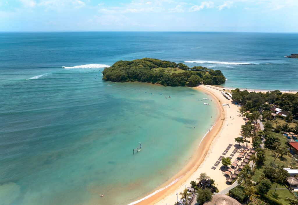 Aerial view of the beach at Paradisus by Meliá Bali in Nusa Dua: a curved sandy bay of calm turquoise water sheltered by a wooded island, with waves breaking further out
