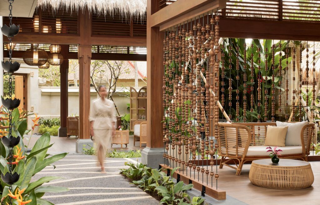 A guest walking along the garden path to YHI Spa at Paradisus by Meliá Bali, past a wooden bead screen and a rattan lounge seat